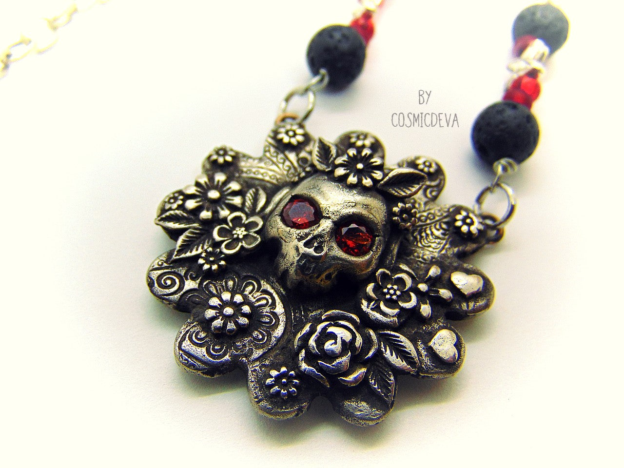 Unique detailed silver bronze Frida Kahlo sugar skull pendant necklace influenced by Mexican culture and symbolism. The flower shaped 3d pendant is featuring a sugar skull decorated with the typical Frida Kahlo signature floral wreath and faceted red garnet cubic zirconia gemstones as eyes. Incorporated flowers, roses, leaves and tiny hearts surrounding the skull. On the backside of the pendant is the CosmicDeva Hallmark