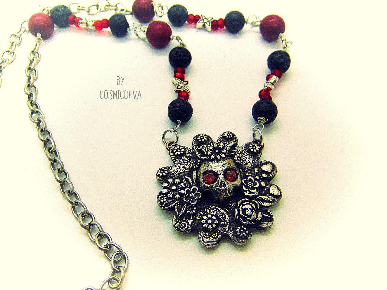 Unique detailed silver bronze Frida Kahlo sugar skull pendant necklace influenced by Mexican culture and symbolism. The flower shaped 3d pendant is featuring a sugar skull decorated with the typical Frida Kahlo signature floral wreath and faceted red garnet cubic zirconia gemstones as eyes. Incorporated flowers, roses, leaves and tiny hearts surrounding the skull. On the backside of the pendant is the CosmicDeva Hallmark