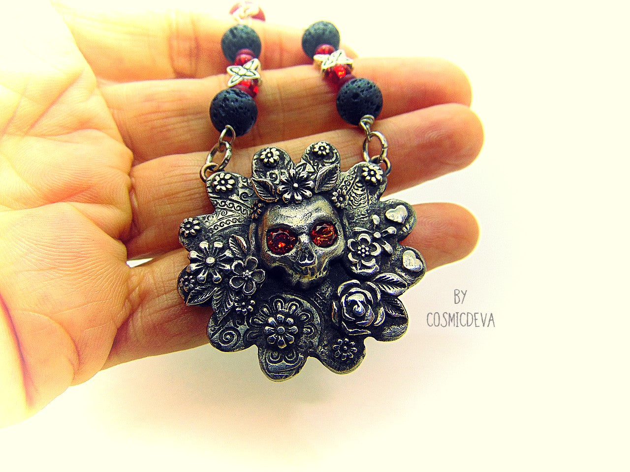 Unique detailed silver bronze Frida Kahlo sugar skull pendant necklace influenced by Mexican culture and symbolism. The flower shaped 3d pendant is featuring a sugar skull decorated with the typical Frida Kahlo signature floral wreath and faceted red garnet cubic zirconia gemstones as eyes. Incorporated flowers, roses, leaves and tiny hearts surrounding the skull. On the backside of the pendant is the CosmicDeva Hallmark