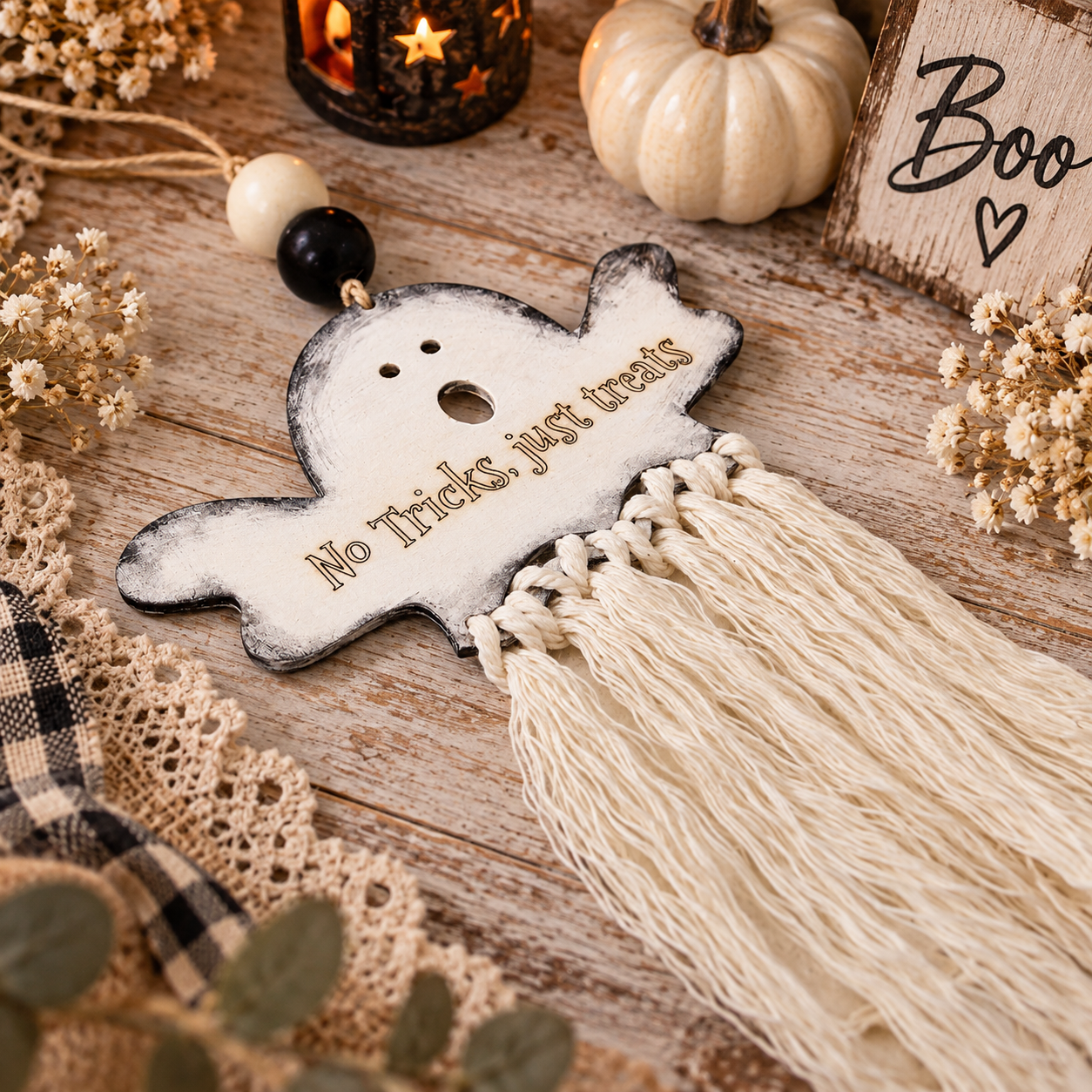 A Halloween-themed macrame tassel ornament shaped like a ghost, featuring the engraved text 'No Tricks, just treats'. The ornament is styled on a rustic wooden table with autumn props including a small pumpkin, lace, dried flowers, and a wooden sign. made by cosmicdeva