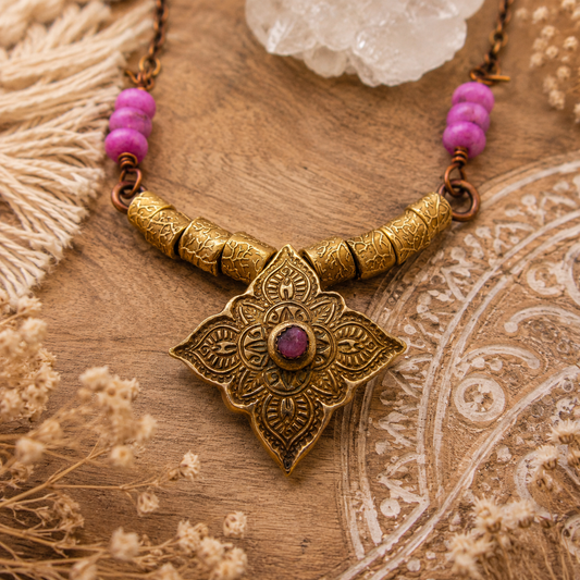 An ornate bohemian-style necklace featuring a large gold-toned floral pendant with a central purple stone, flanked by gold-toned cylindrical beads and purple gemstone beads on a rustic wooden background.