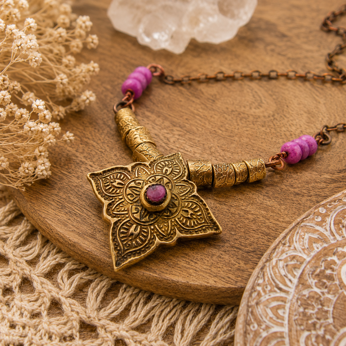 An ornate bohemian-style pendant necklace with a bronze-toned lotus flower pendant, purple beads, and a central purple stone, displayed on a wooden tray with dried flowers and a lace cloth.