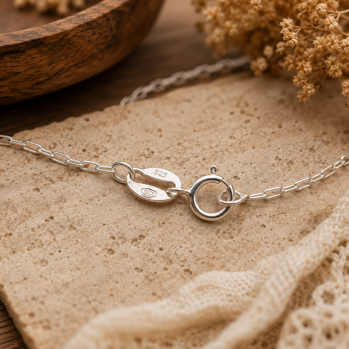 A close-up of a silver chain necklace with a lobster clasp, resting on a textured beige stone tile with dried flowers and a wooden bowl in the background.