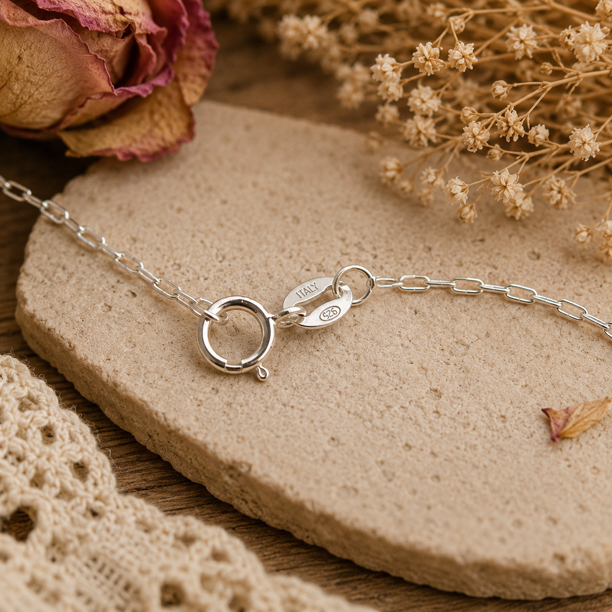A silver chain bracelet with a double-ring clasp and a small engraved tag, styled on a textured beige stone with dried flowers and lace.
