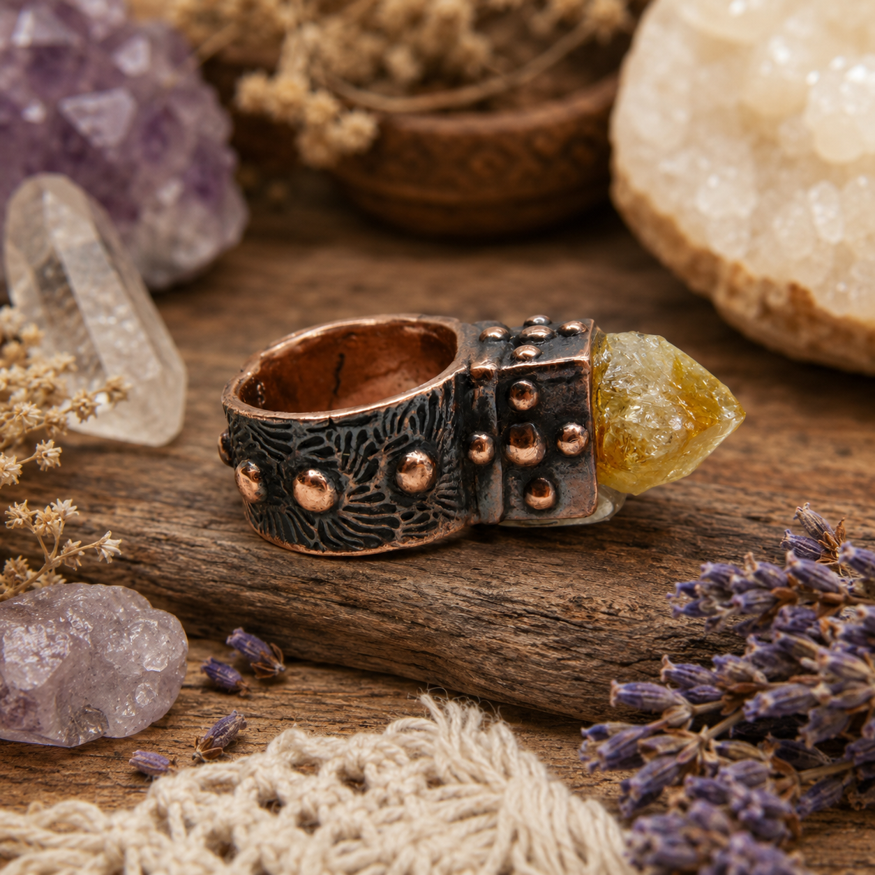 A handmade ring with a textured dark metal band, copper-toned studs, and a yellow-green crystal point, displayed on a rustic wooden surface with crystals and dried flowers.