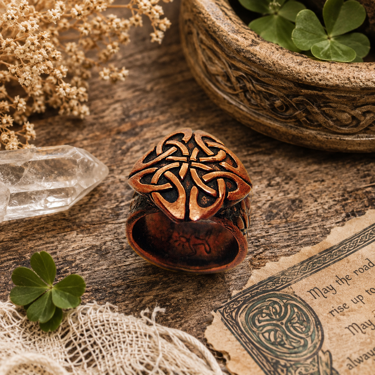 A copper Celtic knot ring with a clover motif, displayed on a rustic wooden surface with dried flowers, a crystal, and a carved bowl. by cosmiceva design 