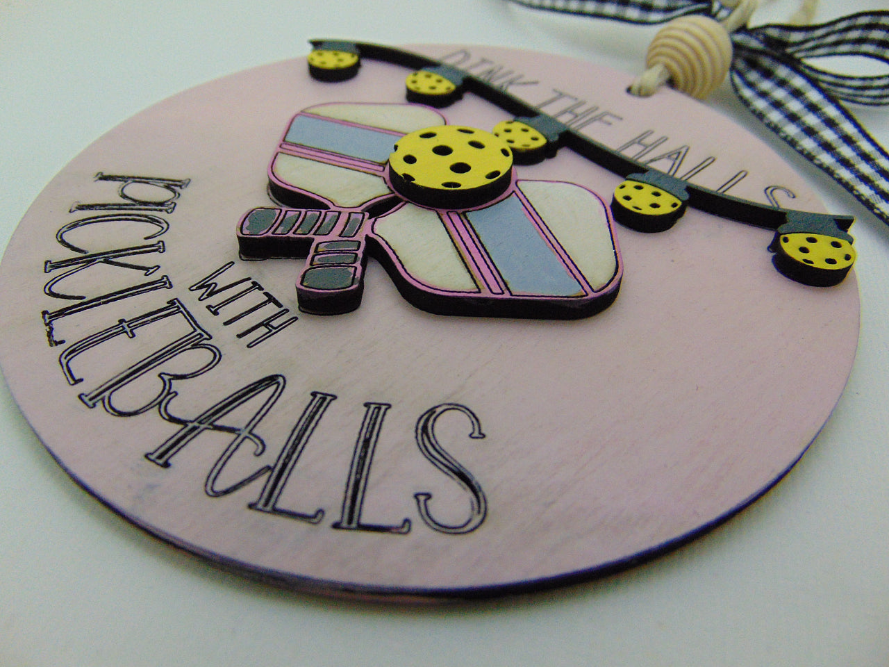 Hand painted pickleball Christmas ornament featuring the engraved phrase “Dink the Halls with Pickleballs.” Multi layered premium wood, 3D design, crossed paddles, festive garland, and handmade checkered bow. A perfect holiday gift for pickleball players and fans.