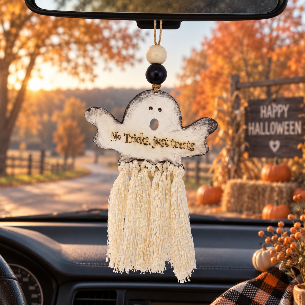 A Halloween-themed car air freshener hangs from the rearview mirror inside a car. The ornament is shaped like a distressed white ghost with the phrase 'No Tricks, just treats' written on it, featuring a cream-colored tassel and decorative beads at the top. made by cosmicdeva