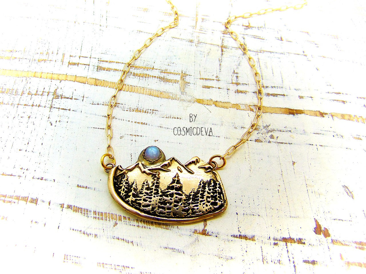 This nature lover landscape pendant necklace features the serenity of a pine forest in the enchanted wilderness of the deep woods with majestic mountains in the background. A flashy rainbow moonstone setting represents a full moon that rises above the top of the mountains. The scenery reminds of the beautiful alpine landscape of Switzerland as well as the great Rocky Mountains.