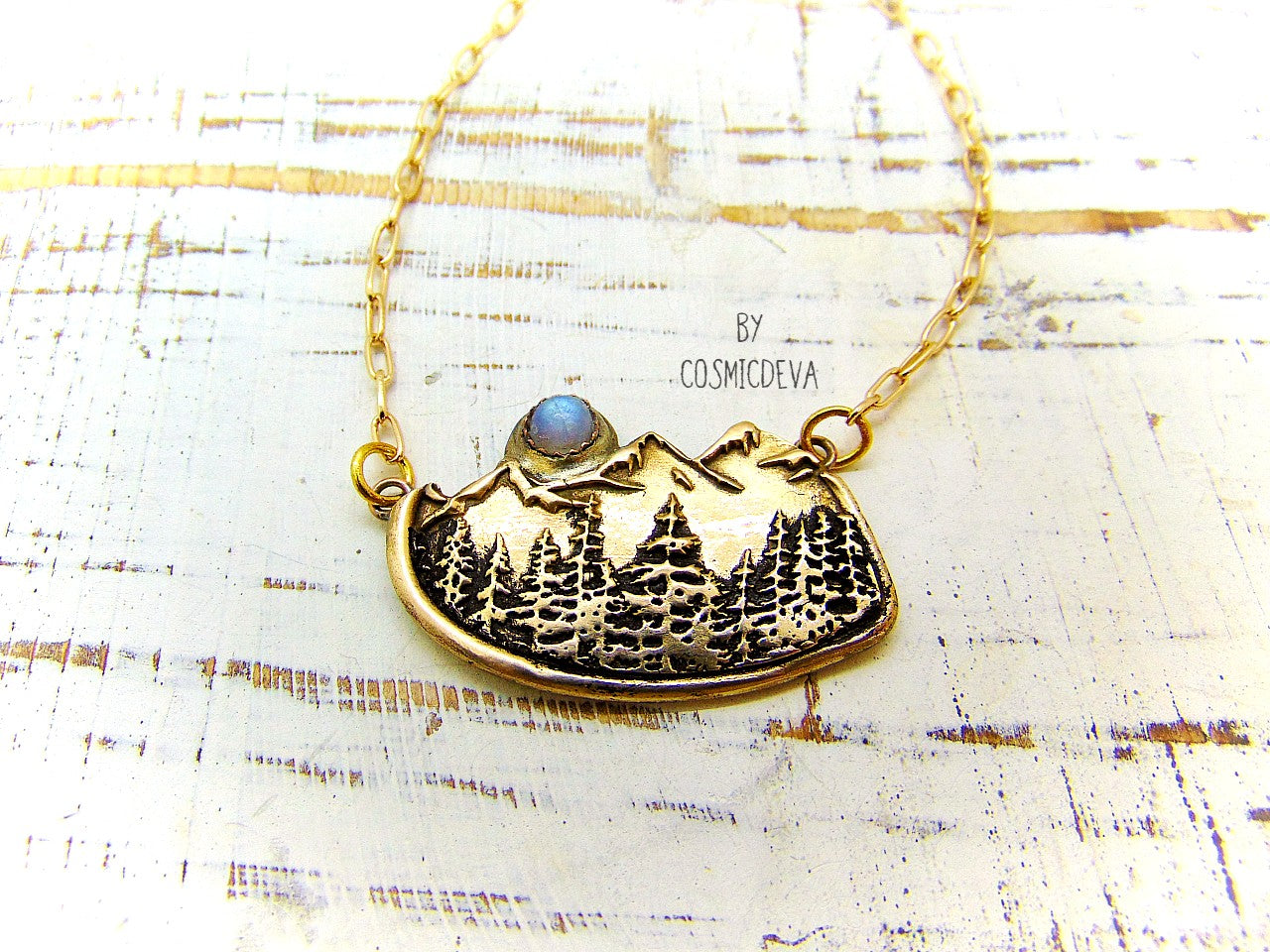 This nature lover landscape pendant necklace features the serenity of a pine forest in the enchanted wilderness of the deep woods with majestic mountains in the background. A flashy rainbow moonstone setting represents a full moon that rises above the top of the mountains. The scenery reminds of the beautiful alpine landscape of Switzerland as well as the great Rocky Mountains.