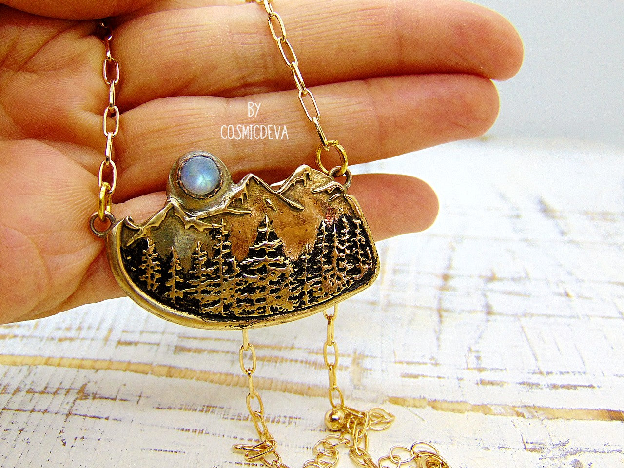 This nature lover landscape pendant necklace features the serenity of a pine forest in the enchanted wilderness of the deep woods with majestic mountains in the background. A flashy rainbow moonstone setting represents a full moon that rises above the top of the mountains. The scenery reminds of the beautiful alpine landscape of Switzerland as well as the great Rocky Mountains.