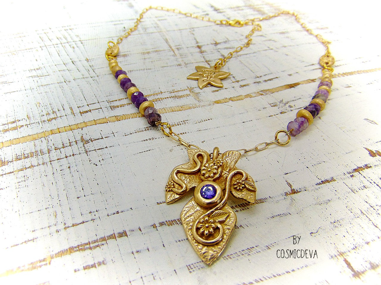 Playful handcrafted fig leaf with an Amethyst in the center and adorned with flowers and swirls. The dainty 18–21-inch necklace is strung with small facetted raw natural lepidolite gemstone rondelle beads and hand formed gold bronze pearls and connectors. I created and casted this leaf with a small fig tree leaf from our garden.